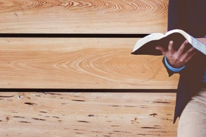 Man leaning against a wood wall reading a Bible